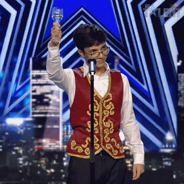a man in a red vest is holding a glass in front of a microphone and the word talent is on the screen behind him