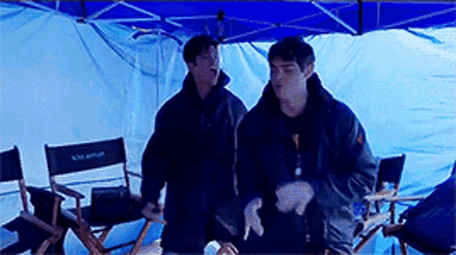 two men standing in front of a blue tent with directors chairs