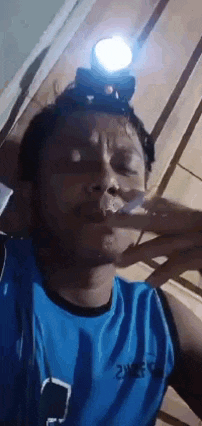 a man in a blue shirt is smoking a cigarette and wearing a headlamp on his head .