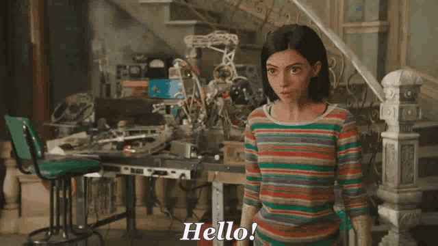 a woman in a colorful striped shirt is standing in a messy room and saying hello .