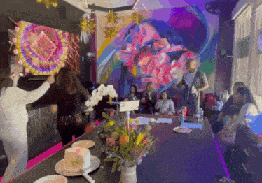 a group of people are gathered around a table in front of a mural of a man 's face