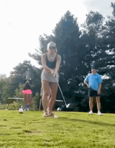 a woman in a skirt is swinging a golf club on a golf course while a man watches .