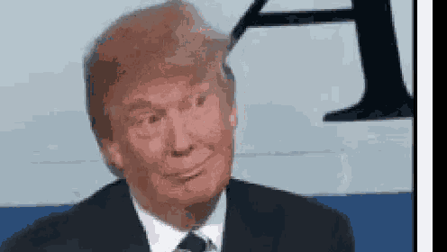 donald trump is wearing a suit and tie and smiling while sitting in front of a sign .