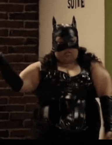 a woman is wearing a cat mask and gloves and standing in front of a brick wall .