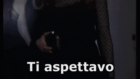 a woman in a black dress is standing in a dark room with the words `` ti aspettavo '' written on the screen .