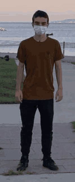 a young man wearing a face mask is standing on a sidewalk near the ocean .