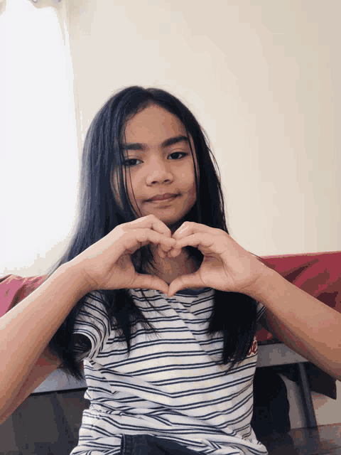 a young girl in a striped shirt makes a heart shape with her hands