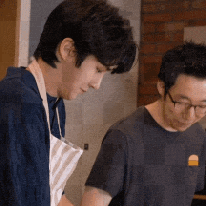 two men are standing next to each other in a kitchen preparing food . one of the men is wearing glasses and an apron .