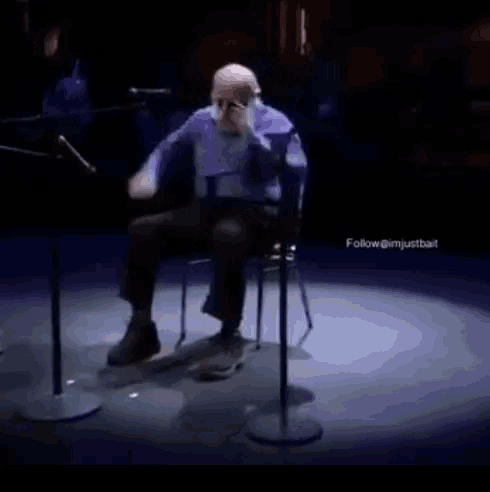 an older man is sitting in a chair on a stage with a microphone in front of him .