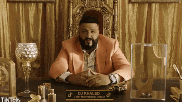 a man in a pink suit is sitting at a desk with a sign that says dj khaled chief motivational officer