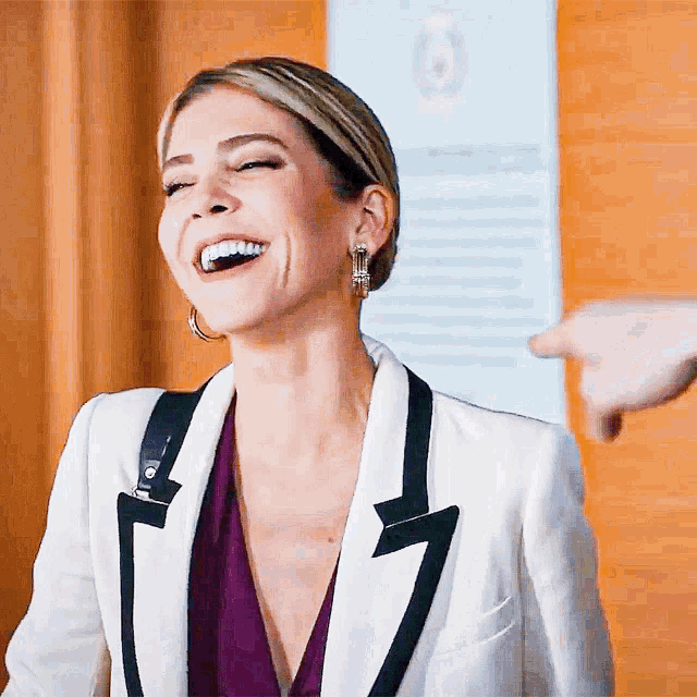 a woman in a white jacket and purple top is smiling and pointing