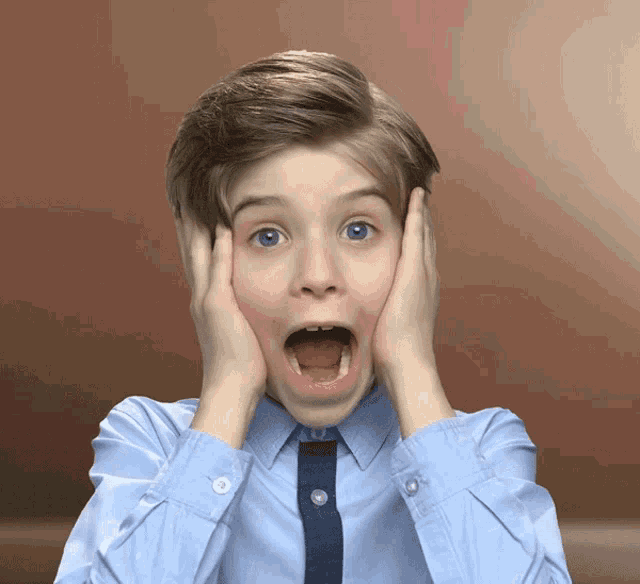 a young boy in a blue shirt and tie has his mouth open and his hands on his face