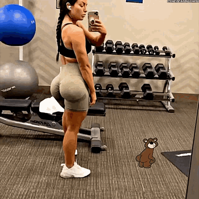 a woman taking a picture of herself in a gym with a teddy bear in the foreground