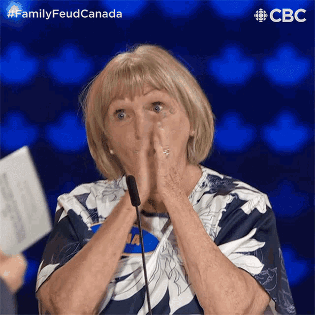 a woman covering her mouth with her hands in front of a microphone with the words family feud canada behind her