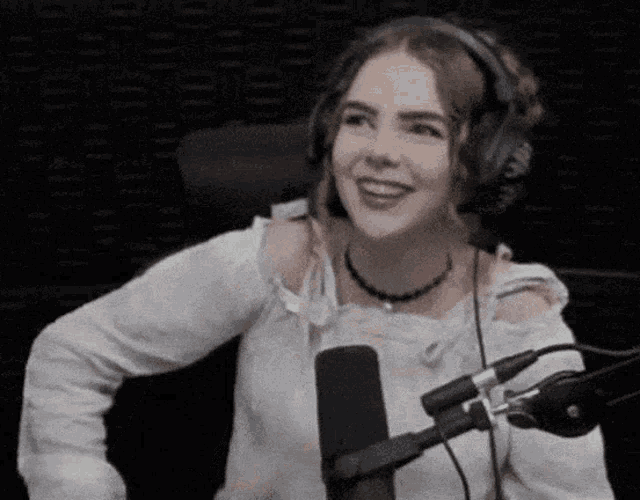 a woman wearing headphones and a white shirt is sitting in front of a microphone and smiling .