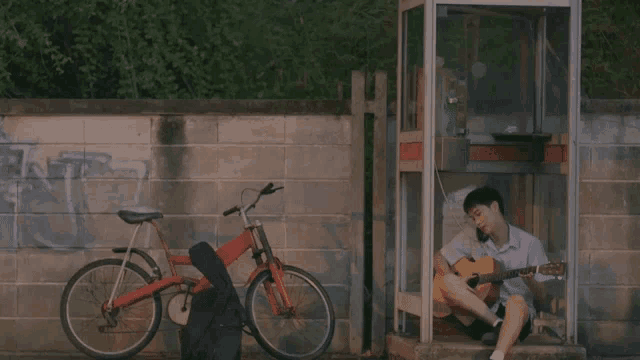 a man sits in a phone booth playing a guitar next to a bike