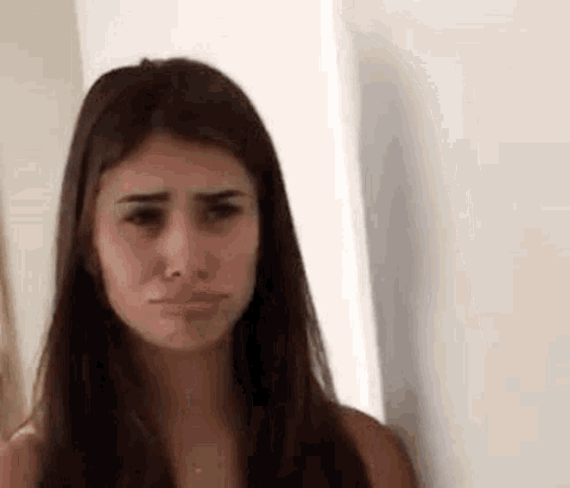 a woman with long brown hair is standing next to a wall and making a sad face .