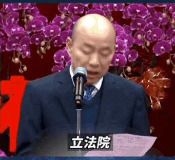 a man in a suit is speaking into a microphone in front of purple flowers
