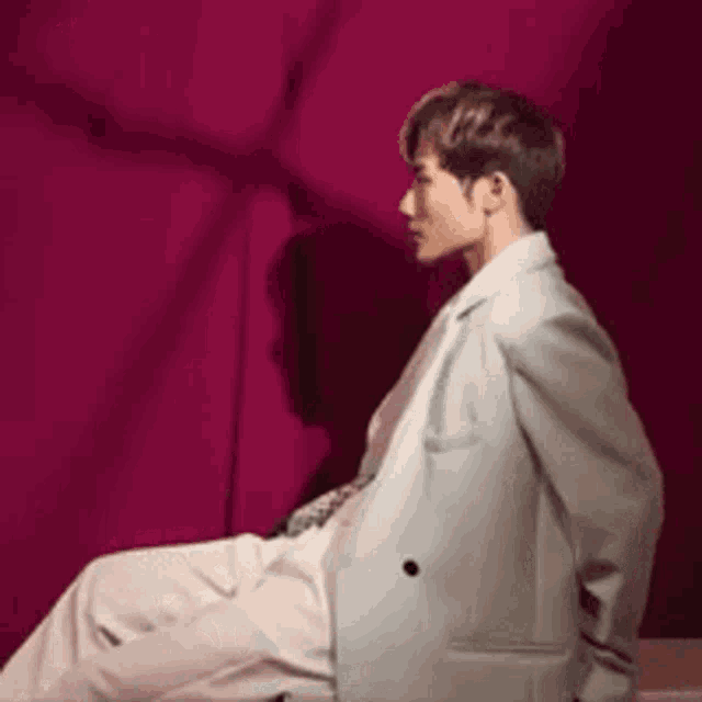 a man in a white suit is sitting in front of a pink curtain .