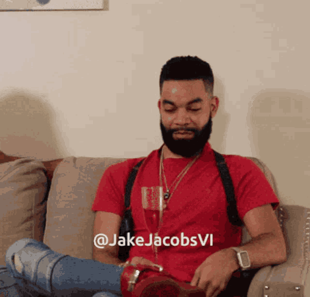 a man with a beard is sitting on a couch with a glass of wine and the hashtag @jakejacobsvi visible