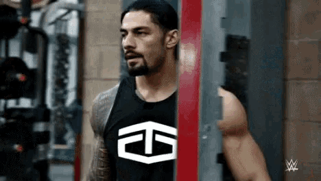 roman reigns is standing in a gym wearing a black tank top with a logo on it .