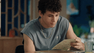 a young man with curly hair is sitting at a table reading a letter