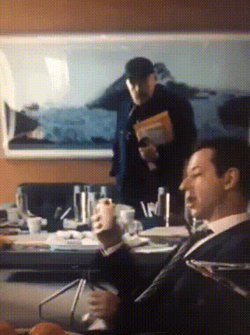 a man in a suit and tie is sitting at a table with a painting in the background