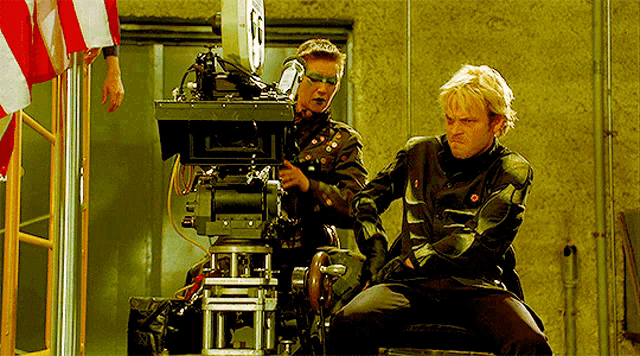 a man is kneeling in front of a camera while another man is standing behind him