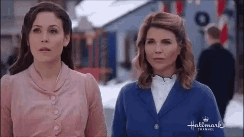 two women are standing next to each other on a snowy street and looking at something .