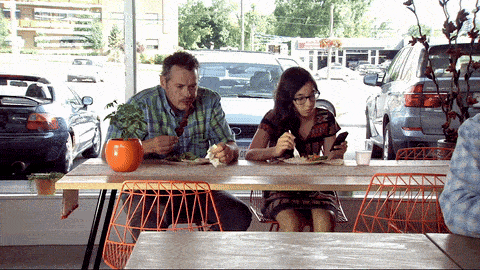 a man and a woman sit at a table in front of a window looking at their phones