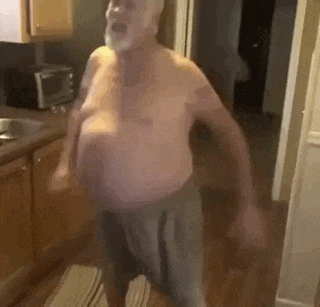an elderly man without a shirt is standing in a kitchen .