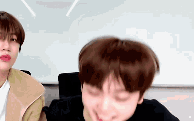 two young men are sitting next to each other in front of a white board and making funny faces .