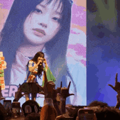 a woman is singing into a microphone in front of a large screen with a picture of her on it .