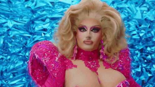 a drag queen is wearing a pink dress and earrings and looking at the camera .