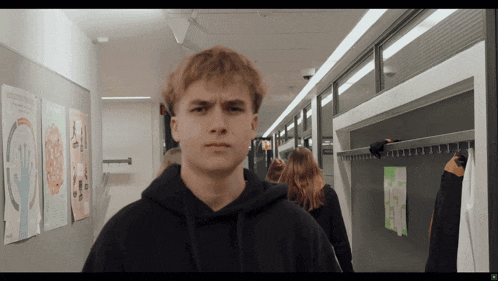 a young man in a black hoodie stands in a hallway with posters on the wall