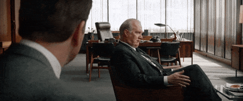 a man in a suit sits in a chair talking to another man in an office