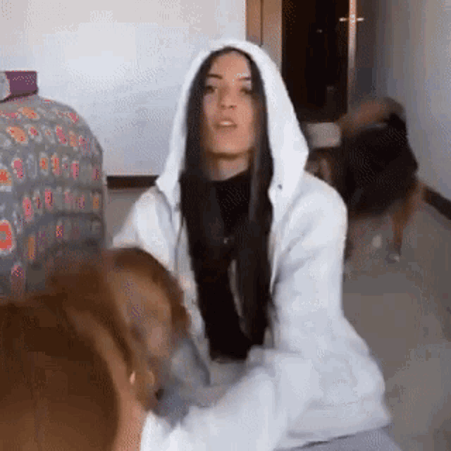 a woman in a white hoodie is playing with a dog in a bedroom .