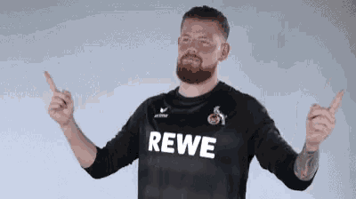 a man with a beard is wearing a black rewe shirt and pointing upwards .