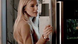 a woman in a brown sweater is standing in front of a refrigerator and smiling