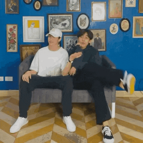 two men are sitting on a couch in front of a wall with pictures .