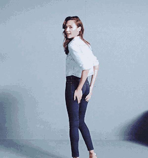 a woman wearing a white shirt and blue jeans is standing in front of a wall