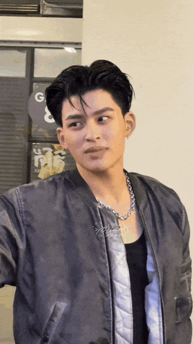 a young man wearing a bomber jacket and a necklace is standing in front of a sign that says g