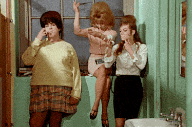 three women standing next to each other in a bathroom with one smoking a cigarette