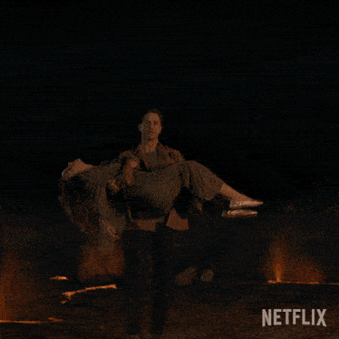 a man is carrying a woman in front of a fire with netflix written on the bottom right