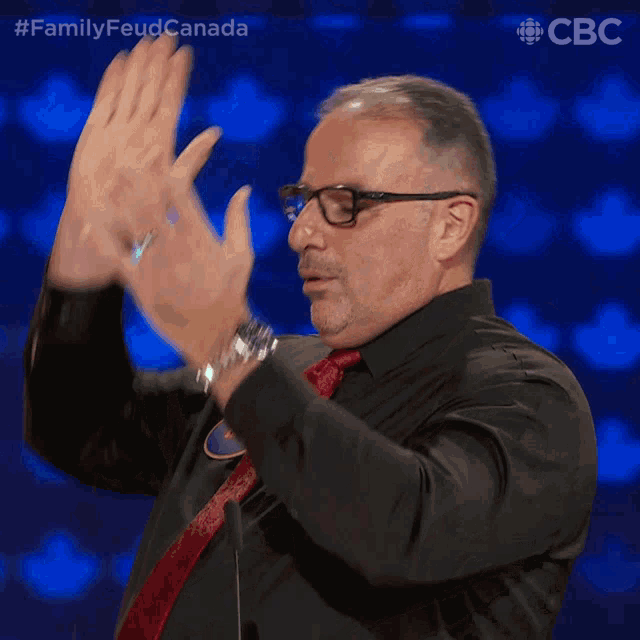 a man wearing glasses and a black shirt applauds in front of a blue background with the letters cbc on it