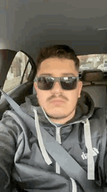 a man wearing sunglasses and a seat belt is taking a selfie while sitting in a car .