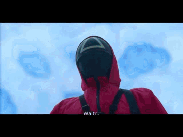 a man in a red jacket and mask is standing in front of a blue background and talking .