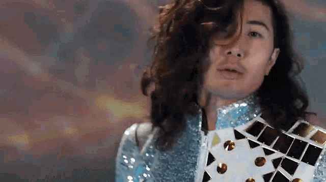 a man with long curly hair is wearing a silver jacket with gold studs