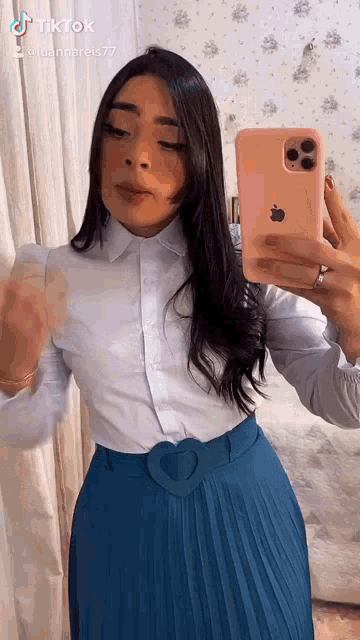 a woman is taking a selfie with her phone while wearing a white shirt and blue pleated skirt .