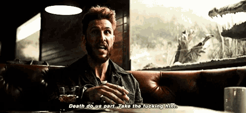 a man with a beard is sitting in a booth with a glass of whiskey and says death do us part